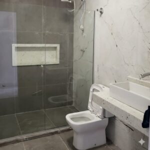 Imagem de um banheiro com vaso sanitário e lavabo brancos, parede com textura de marmorino e box com blindex.